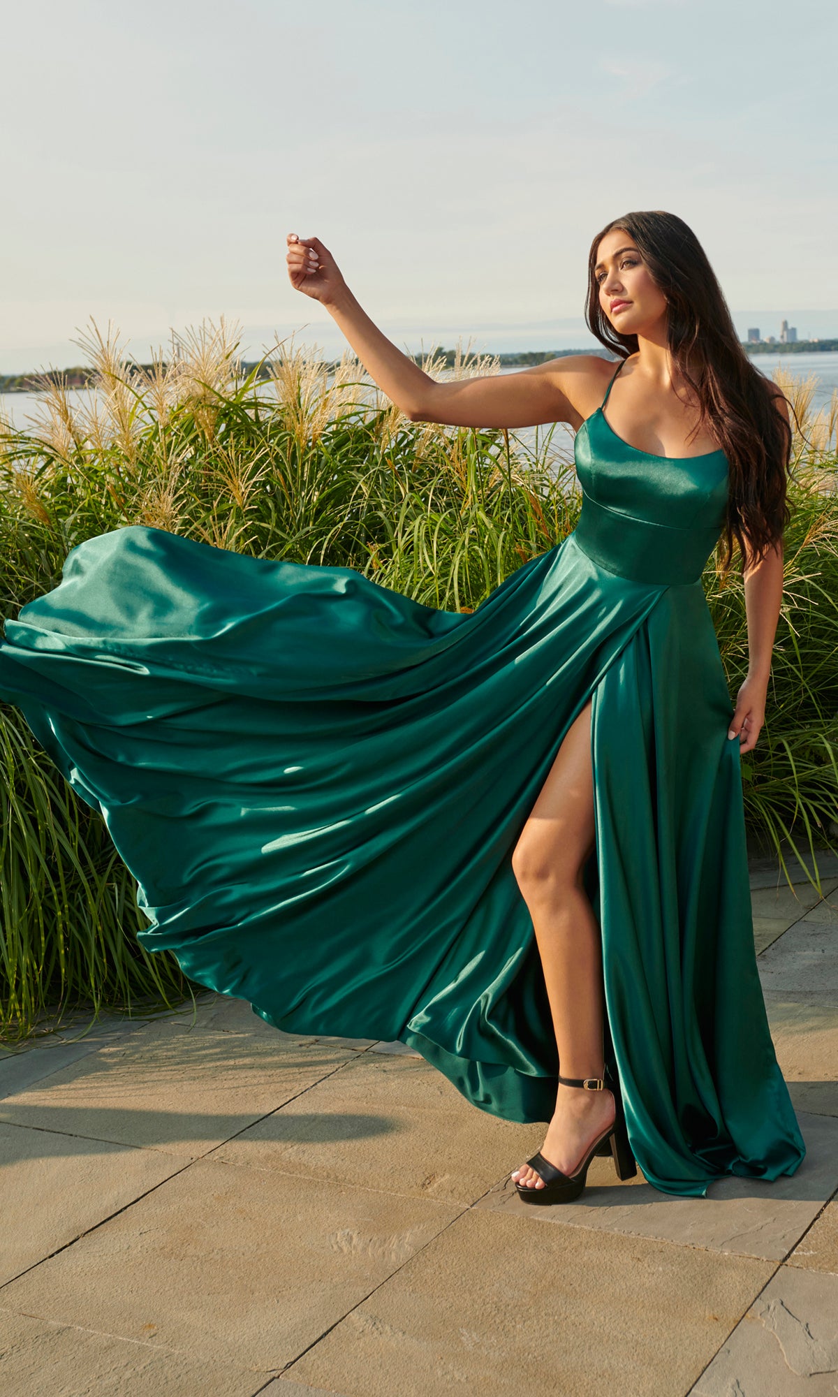 A woman stands outdoors near tall grass, holding out part of her Long Open-Back Formal Prom Dress by Faviana—an elegant emerald green gown with a thigh-high slit.